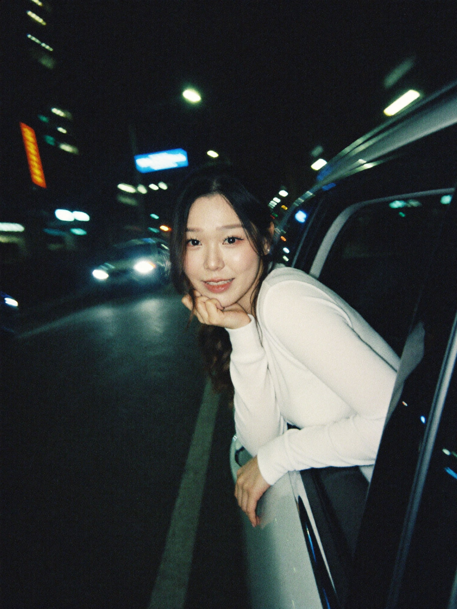 Dreamy car window portrait: a woman in a white long-sleeved top leaning out of a car window at night, cradling her chin, motion-blurred city lights behind