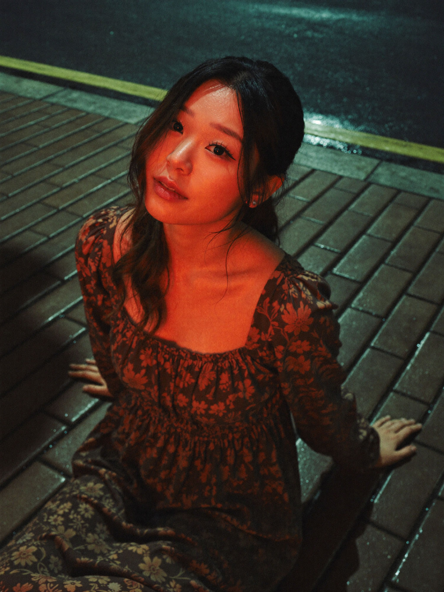 Wet pavement night portrait: a woman in a dark floral square-neck dress seated on wet brick under intense red and orange light