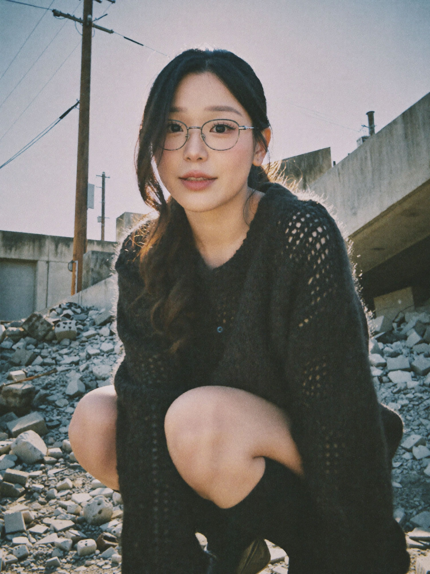 Sunlit urban ruins portrait: a woman in glasses and a fuzzy black knit sweater crouching among concrete rubble, sun haloing her hair