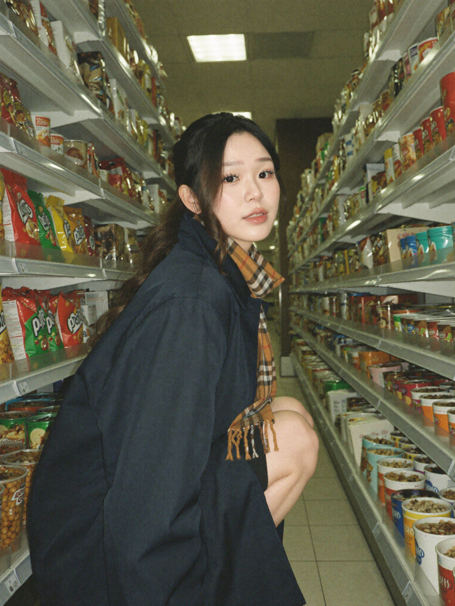 Convenience store portrait: a woman in an oversized navy coat and plaid scarf crouching between packed snack aisles under fluorescent light