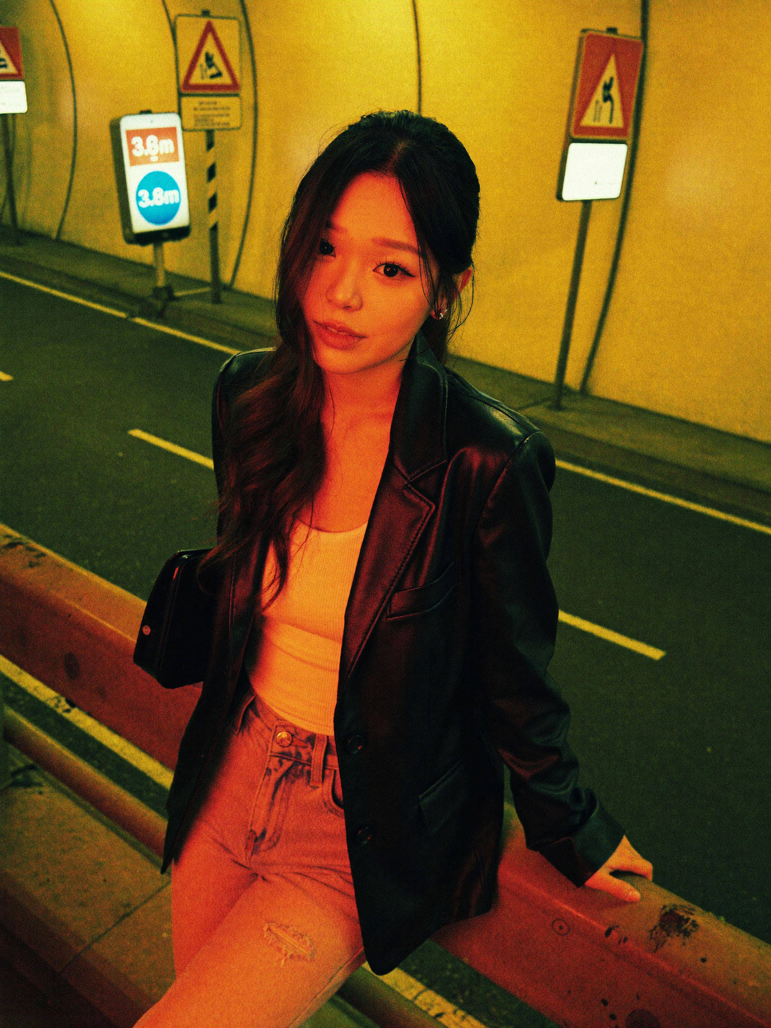 Neo-noir tunnel portrait: a woman in a black leather blazer leaning on a guardrail inside a night tunnel, lit by orange and cold green cross-light