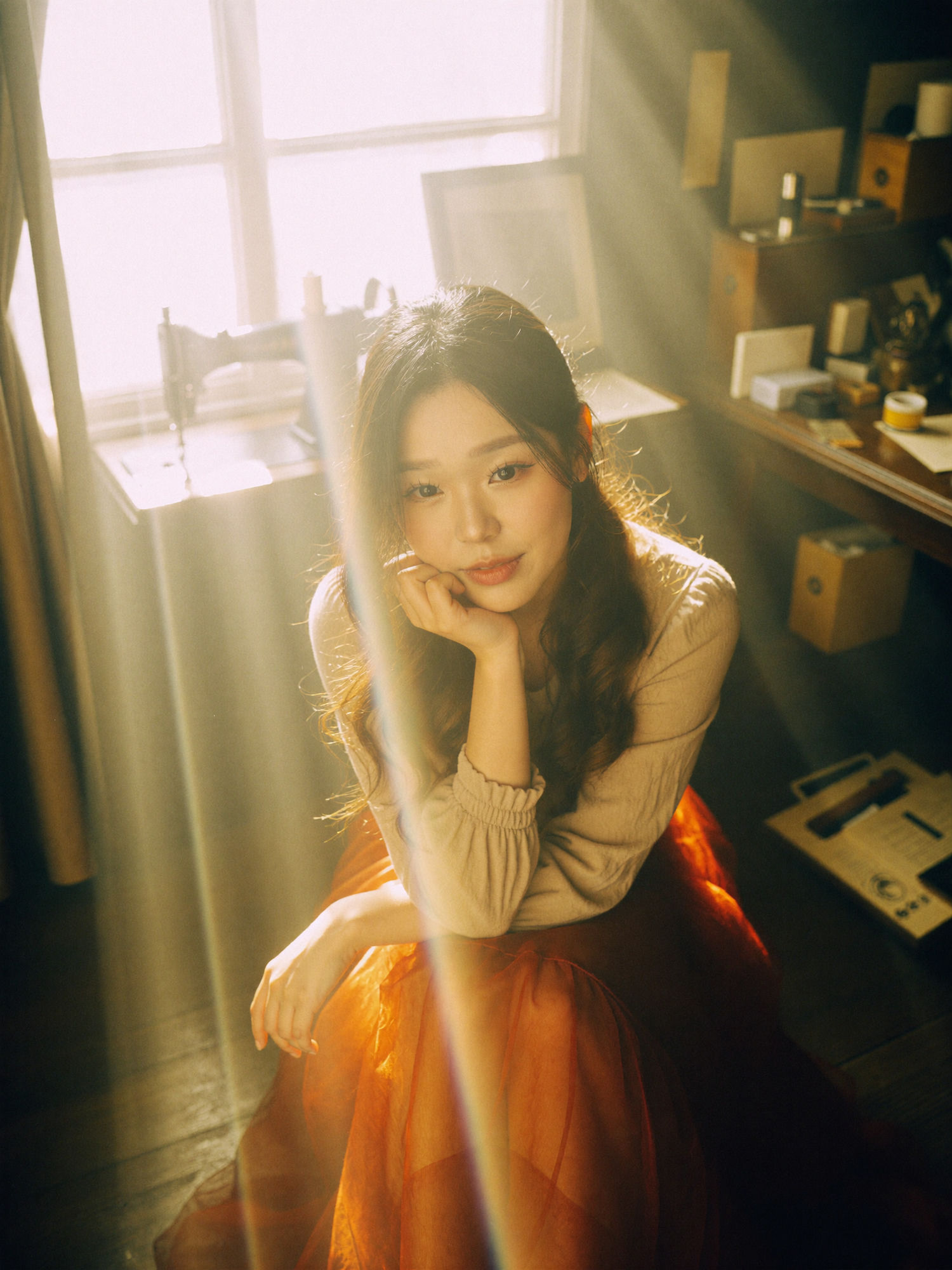 Sunlit window portrait: a woman in a beige top and sheer red-orange skirt seated by a bright window with a vintage sewing machine, golden light flares