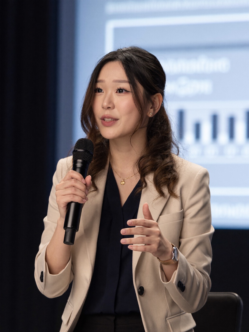 Beige blazer and navy blouse, speaking on stage with a microphone